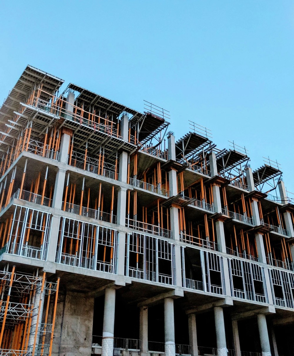 A multi-storey concrete building frame under construction, with orange props and steel stud framing against a clear blue sky.